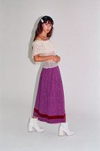 Woman wearing a white lace top and purple skirt with white boots on a plain background