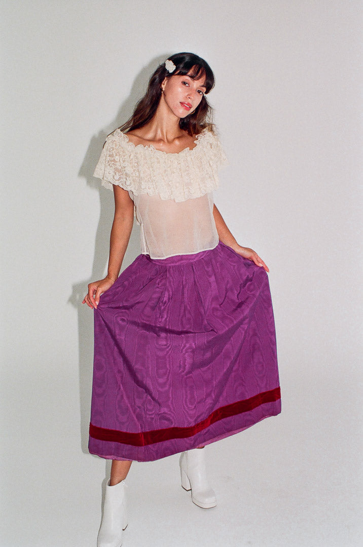 Woman wearing a white top with floral details and a purple skirt on a plain background