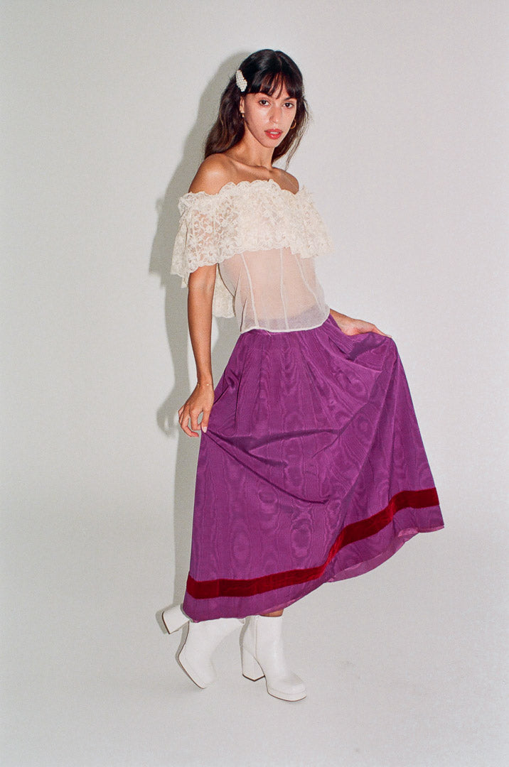 Woman wearing a white off-shoulder top and purple skirt with red trim on a plain background