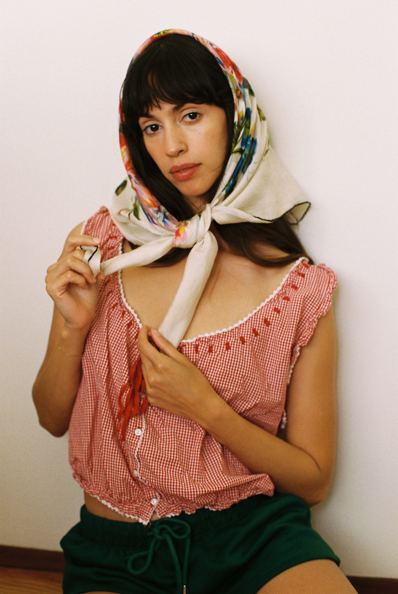 Woman wearing a red checkered top and green shorts with a colorful headscarf against a white background