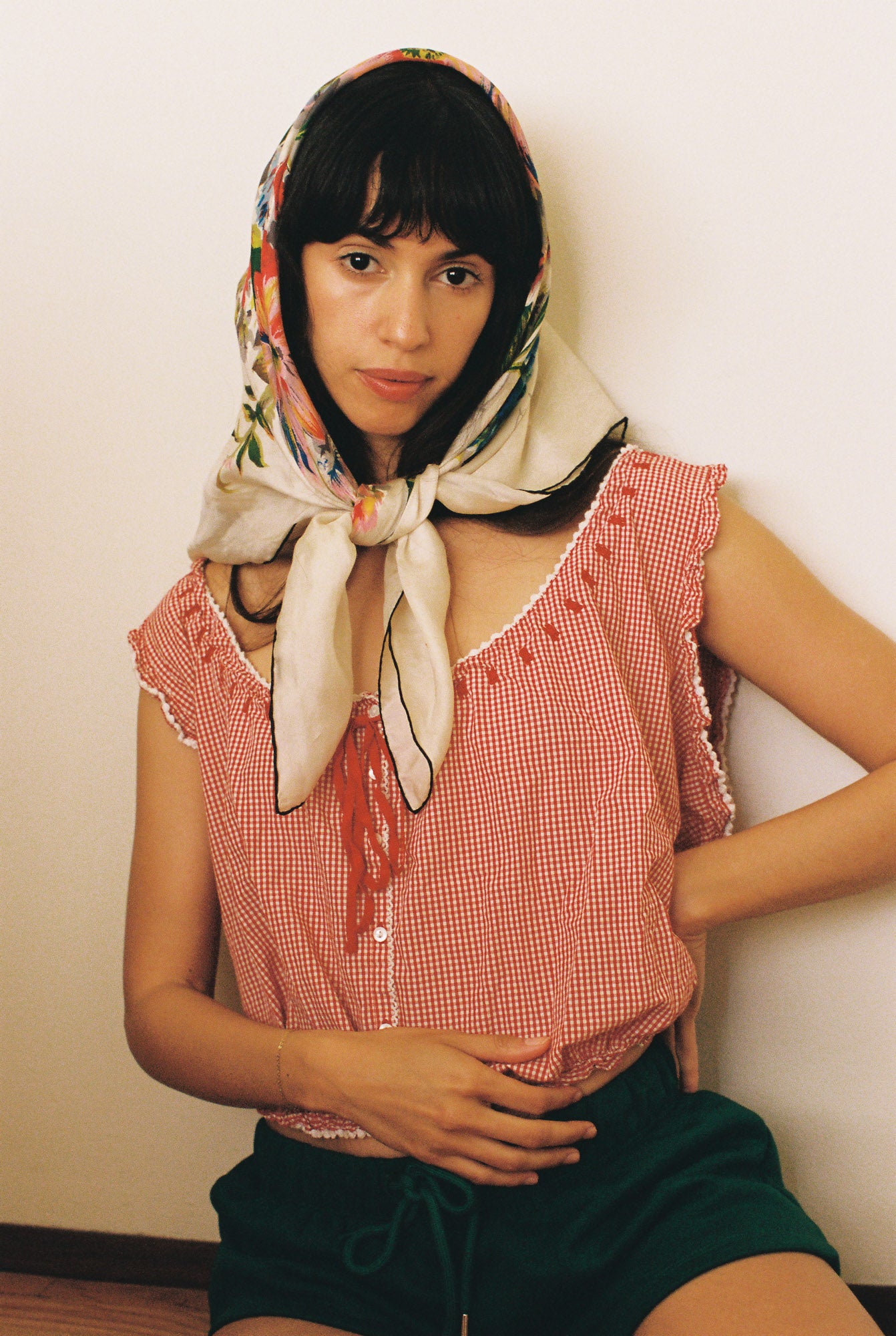 Woman wearing a red checkered top and green shorts with a colorful headscarf against a plain background