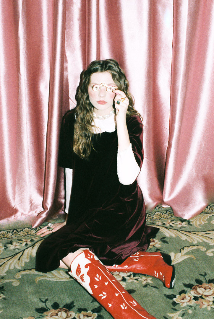 Person sitting on the floor against a pink curtain wearing a black dress and red boots with white patterns.