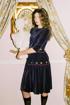 Woman wearing a dark blue velvet dress with floral embroidery in front of an ornate mirror.