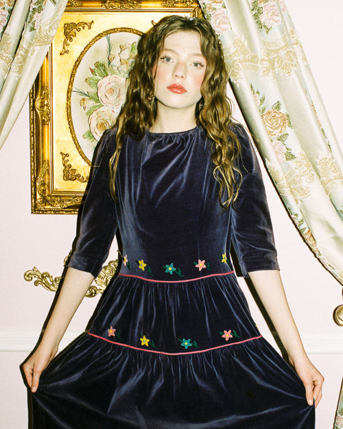 Woman wearing a dark blue velvet dress with floral embroidery in a decorative interior setting.