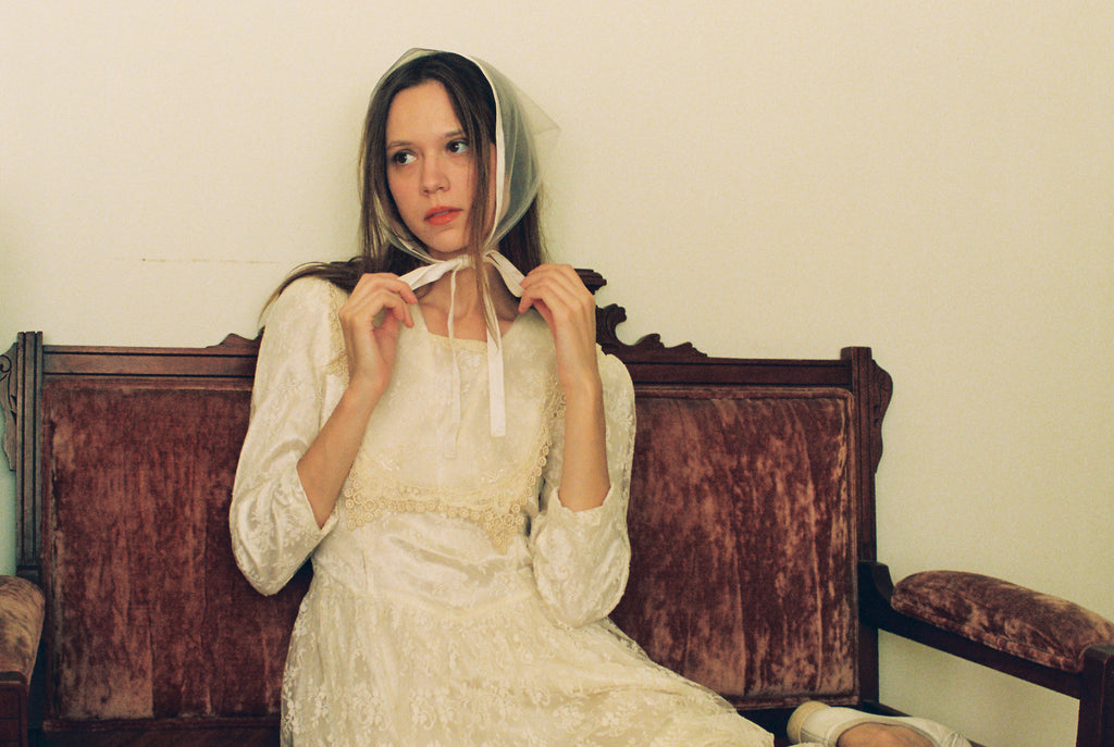 Woman in a white dress sitting on an old wooden bench against a plain wall.