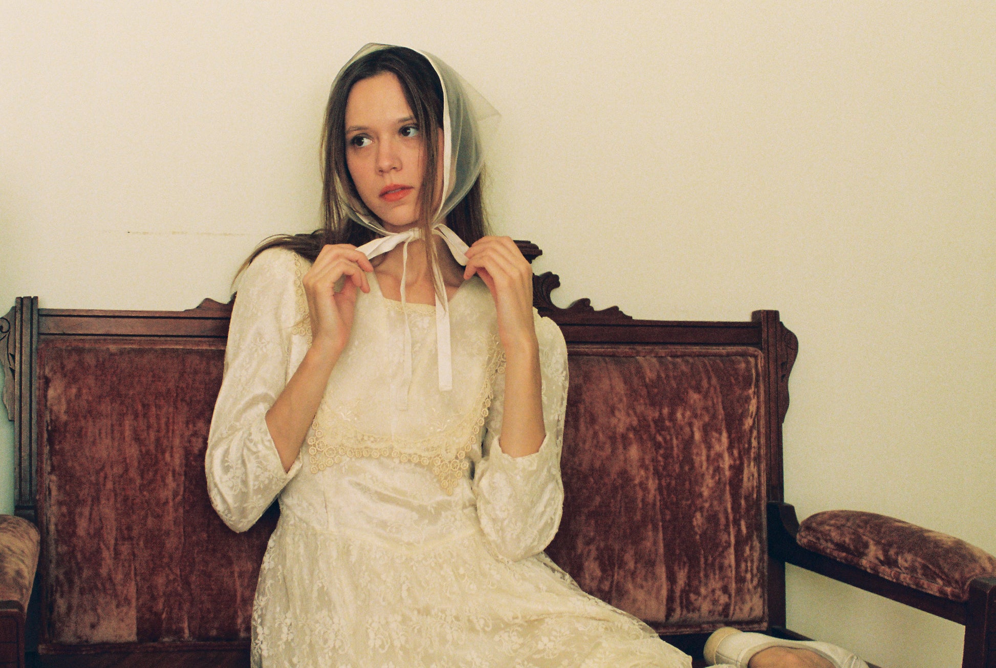 Woman in a white dress sitting on an old wooden bench against a plain wall.