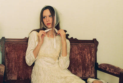 Woman in a white dress sitting on an old wooden bench against a plain wall.