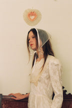 Woman wearing a lace headscarf and white dress against a plain wall.