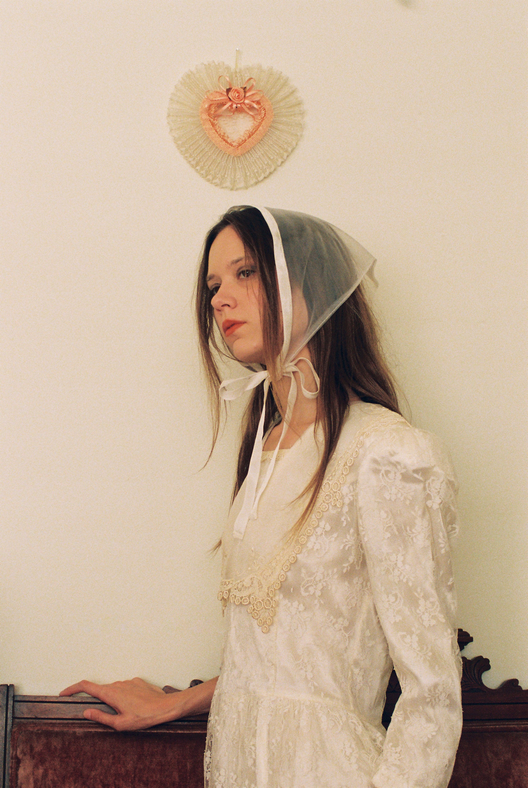 Woman wearing a lace headscarf and white dress against a plain wall.
