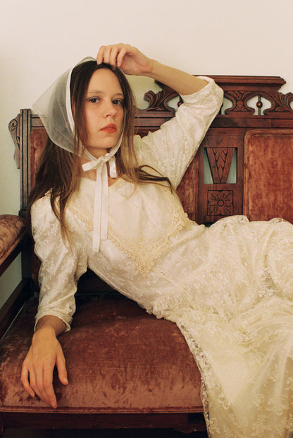Woman in a white lace dress and bonnet sitting on an ornate wooden bench.