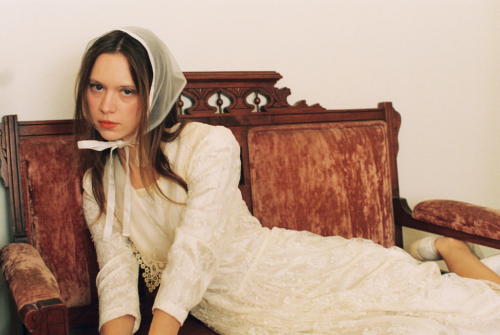 Woman in a white dress and headscarf sitting on an ornate wooden bed.