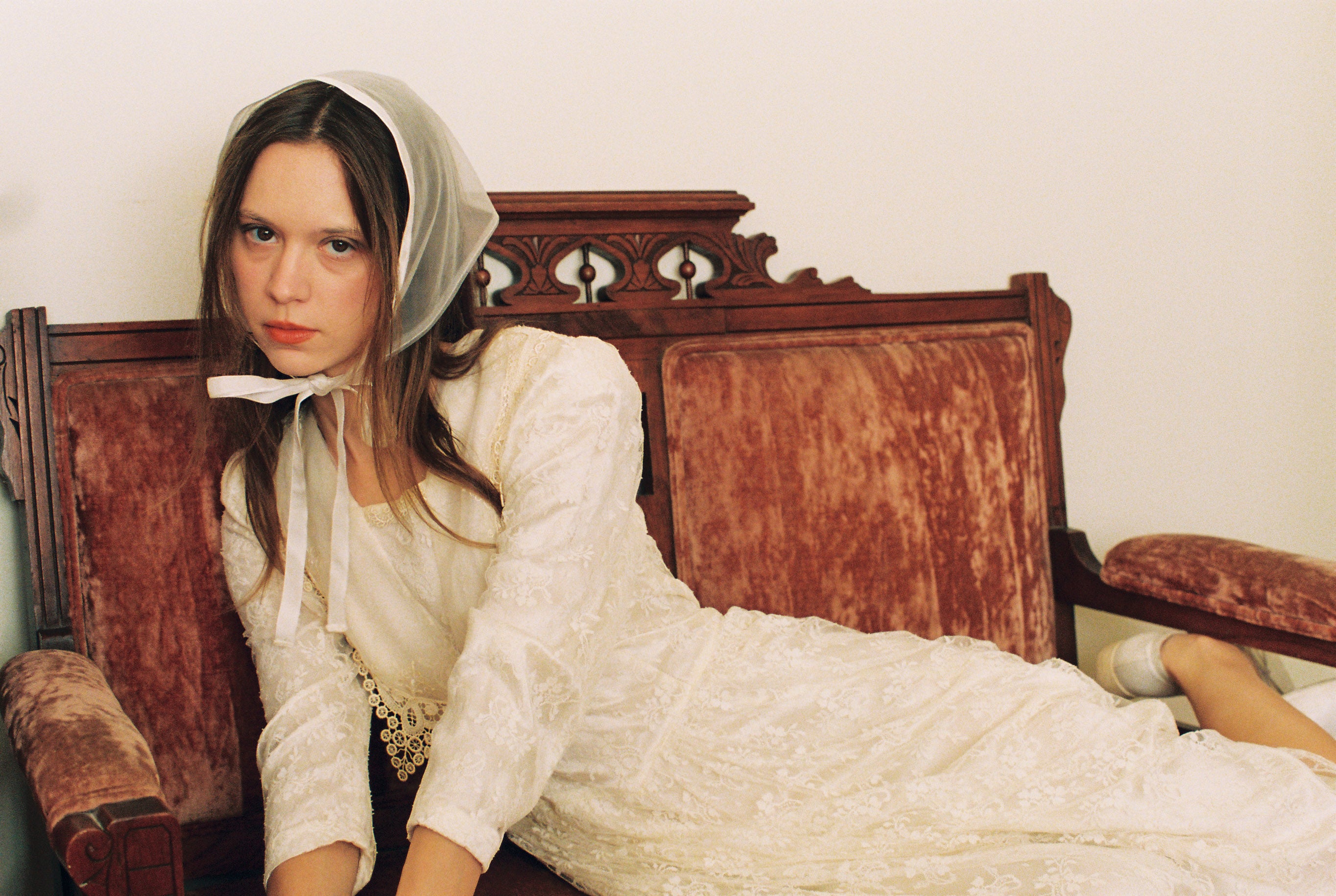 Woman in a white dress and headscarf sitting on an ornate wooden bed.