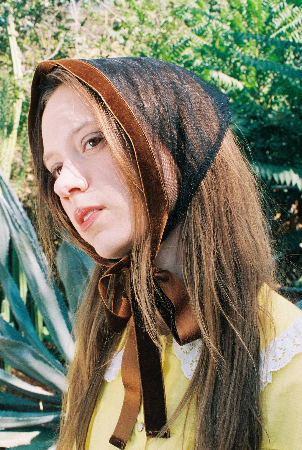 Woman wearing a brown headscarf with a yellow top in front of green foliage