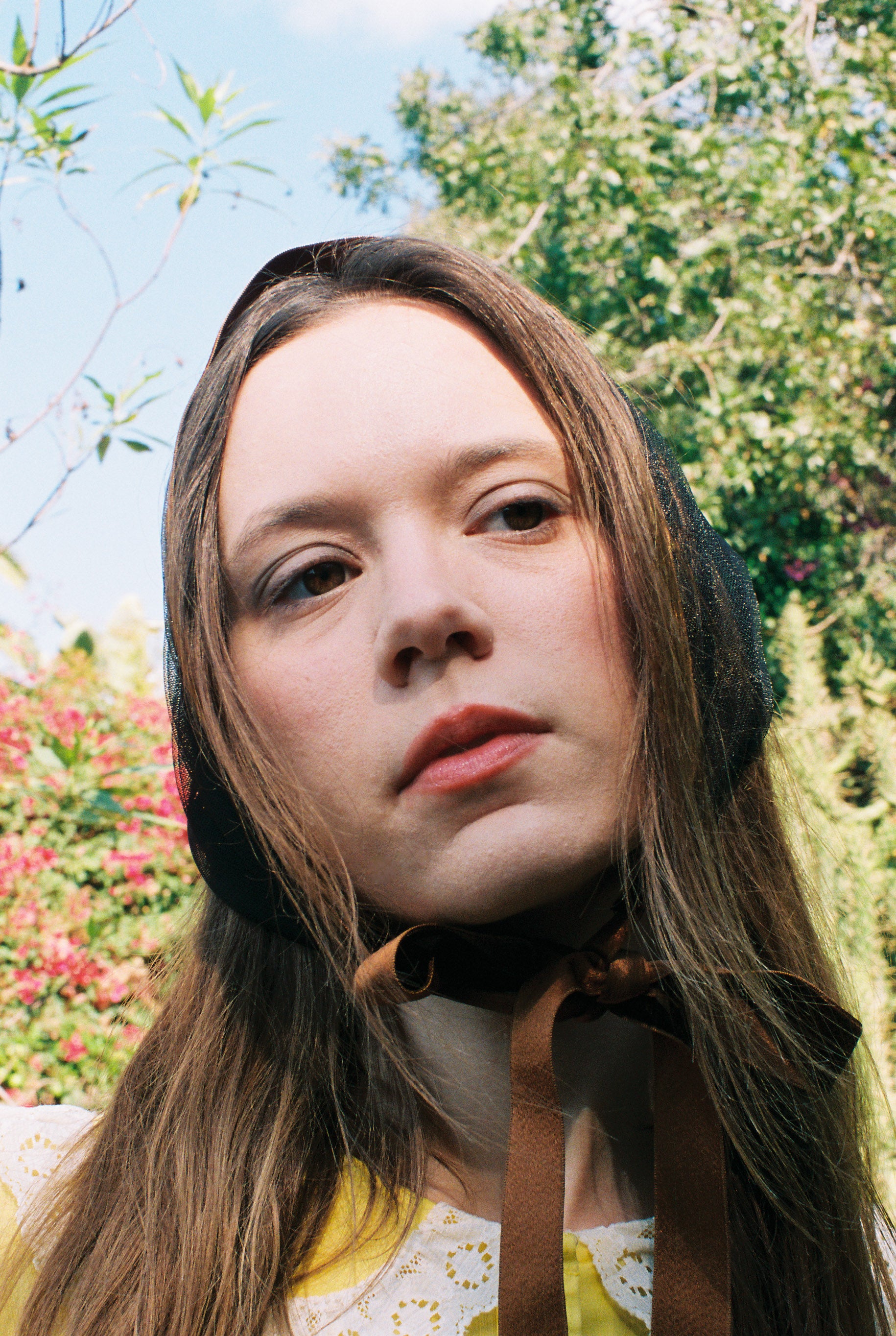 Person with long hair and a bow tie standing outdoors with trees and flowers in the background