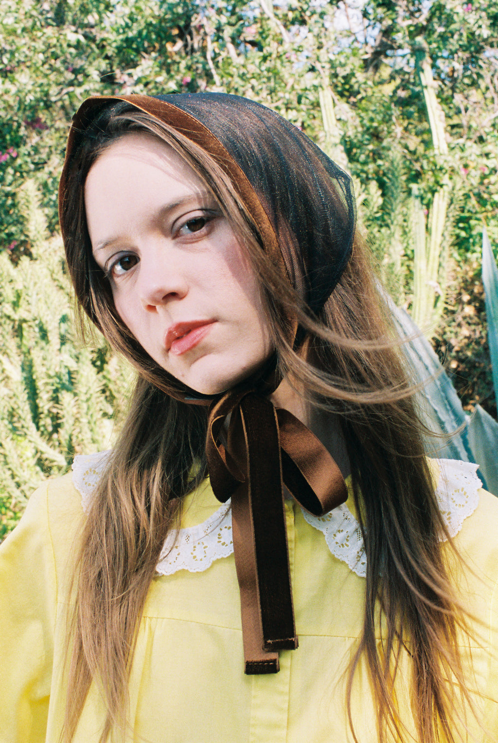 Person wearing a brown headscarf with a bow, standing outdoors with greenery in the background