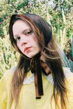 Person wearing a brown headscarf with a bow, standing outdoors with greenery in the background