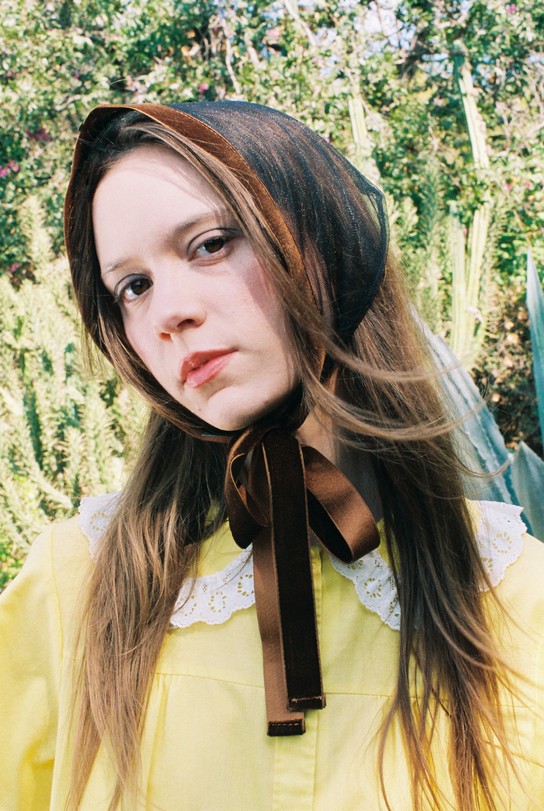 Person wearing a brown headscarf with a bow, standing outdoors with greenery in the background